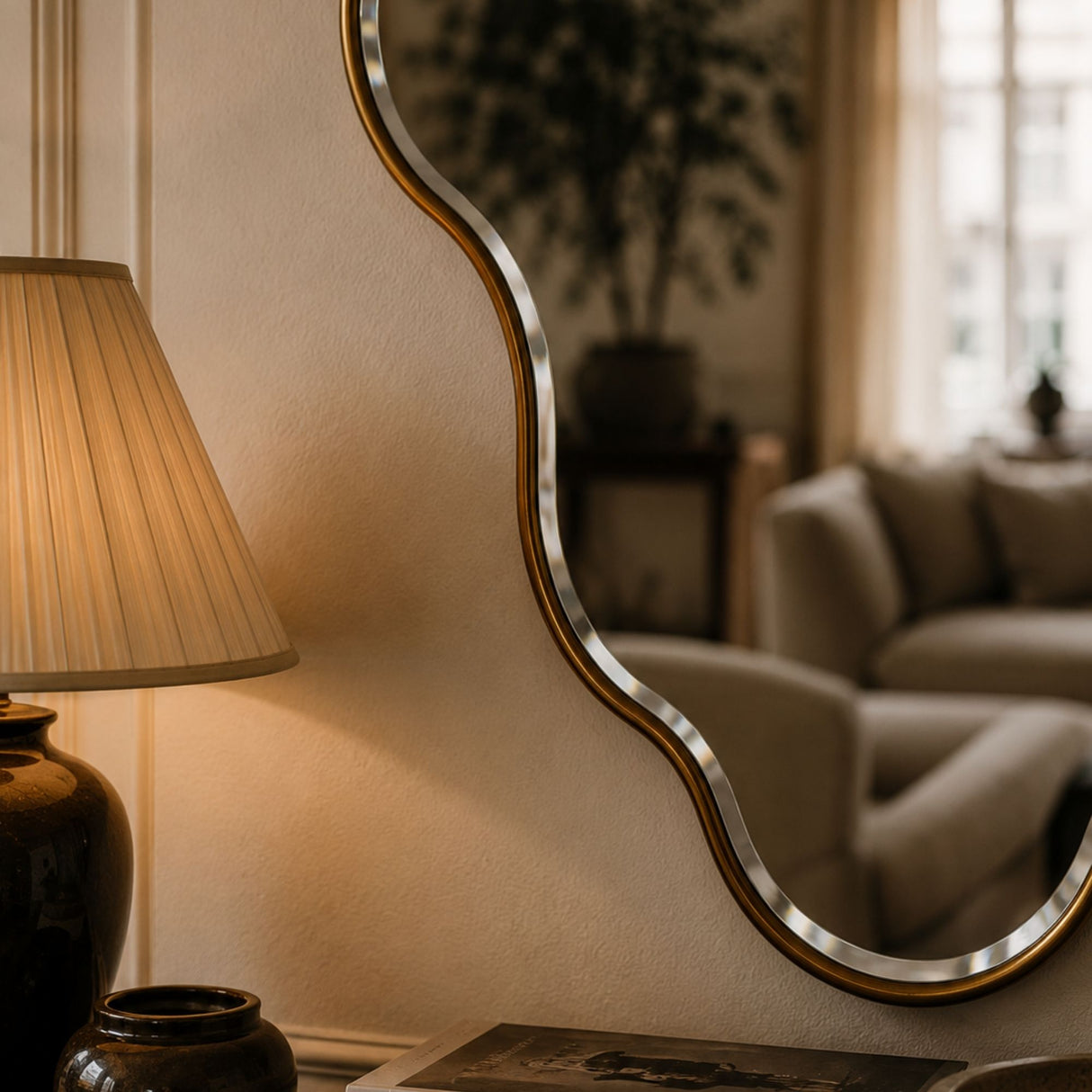 A wavy, organic-shaped wall mirror with a brushed silver metal frame and a gold-toned inner border, styled in a warm, cozy living room next to a lit table lamp.