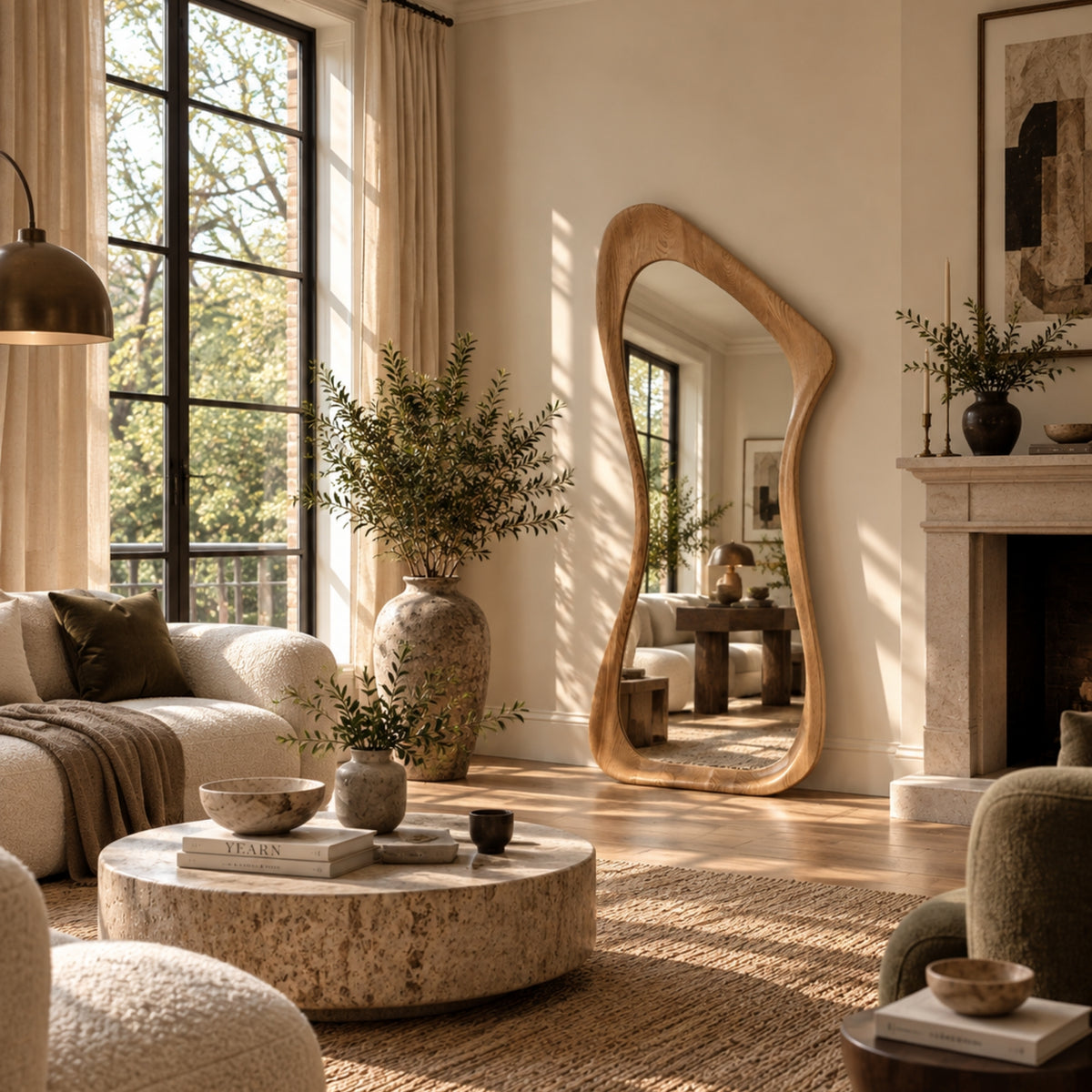 A large, organic-shaped wooden floor mirror with a natural wood frame stands in a bright, modern living room with beige walls, a stone coffee table, and large potted plants.