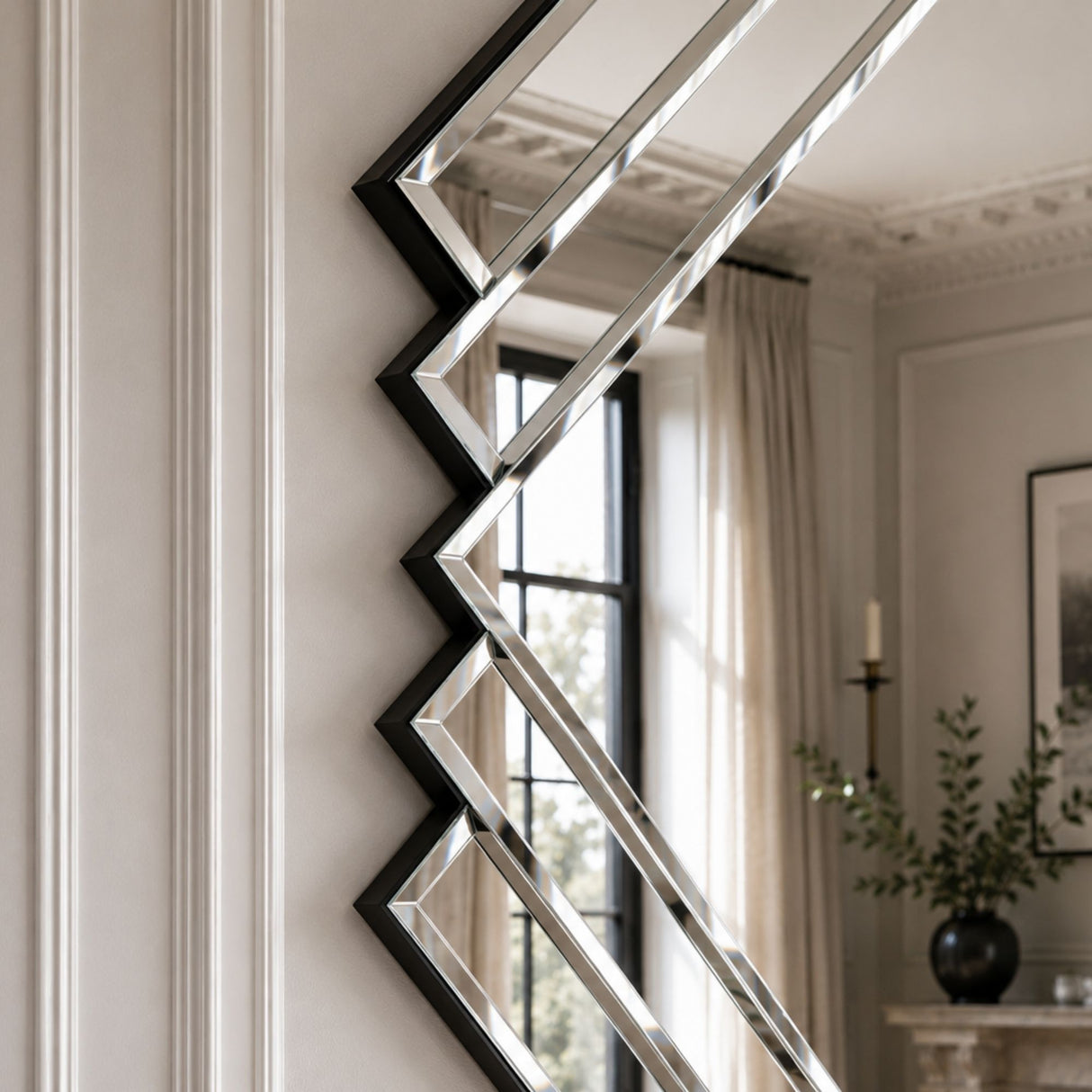 A modern geometric wall mirror with a black zigzag frame and reflective silver mirrored panels, mounted on a white paneled wall in a styled interior room.