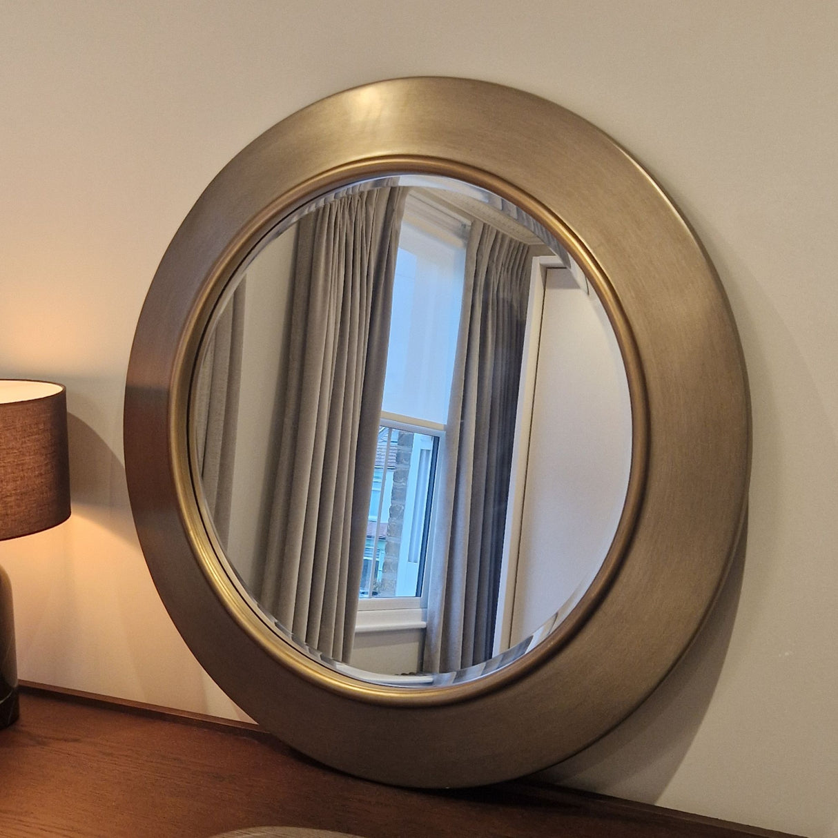 Round metallic mirror on a wall with a lamp and window reflection.