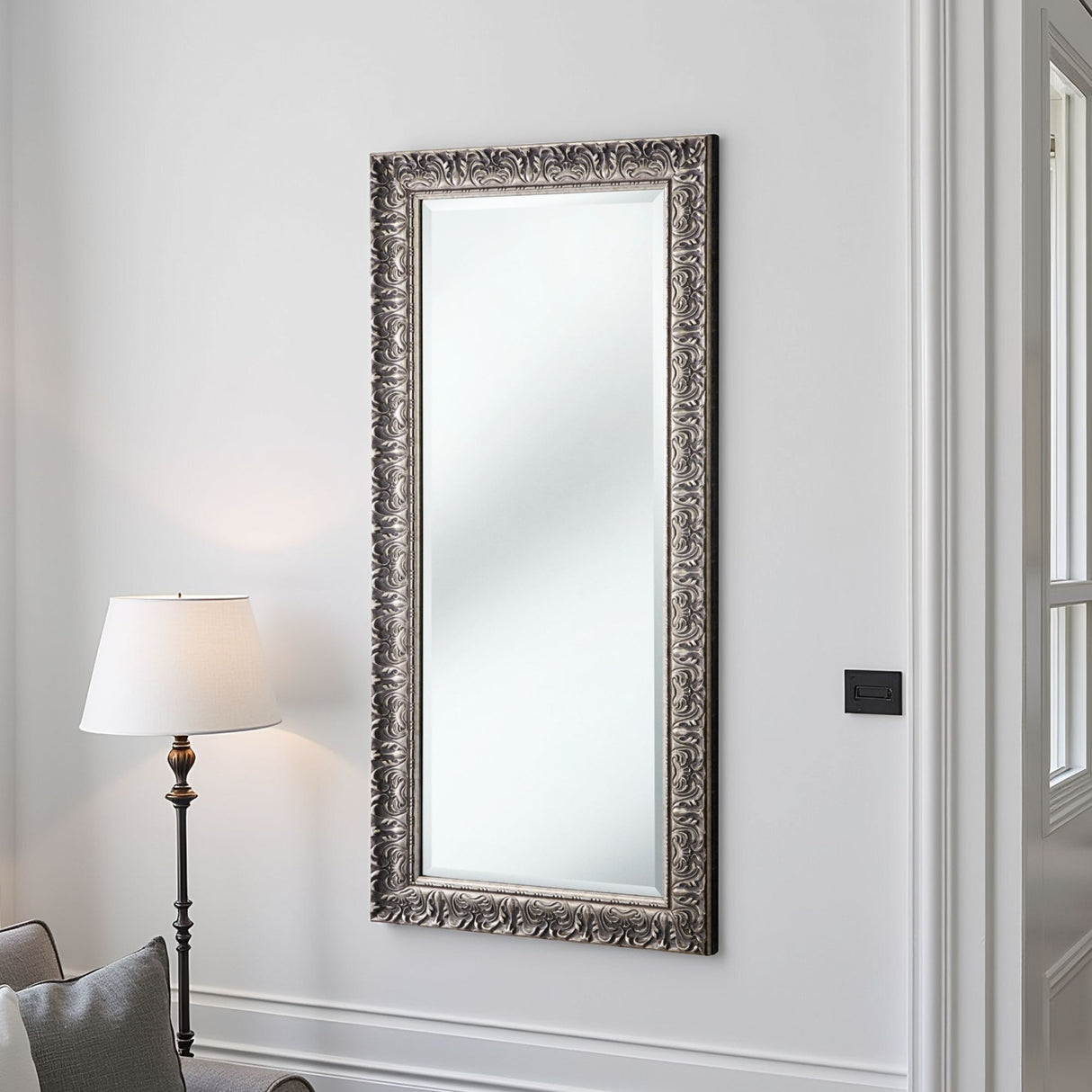 Decorative wall mirror with ornate frame on a white wall next to a lamp.