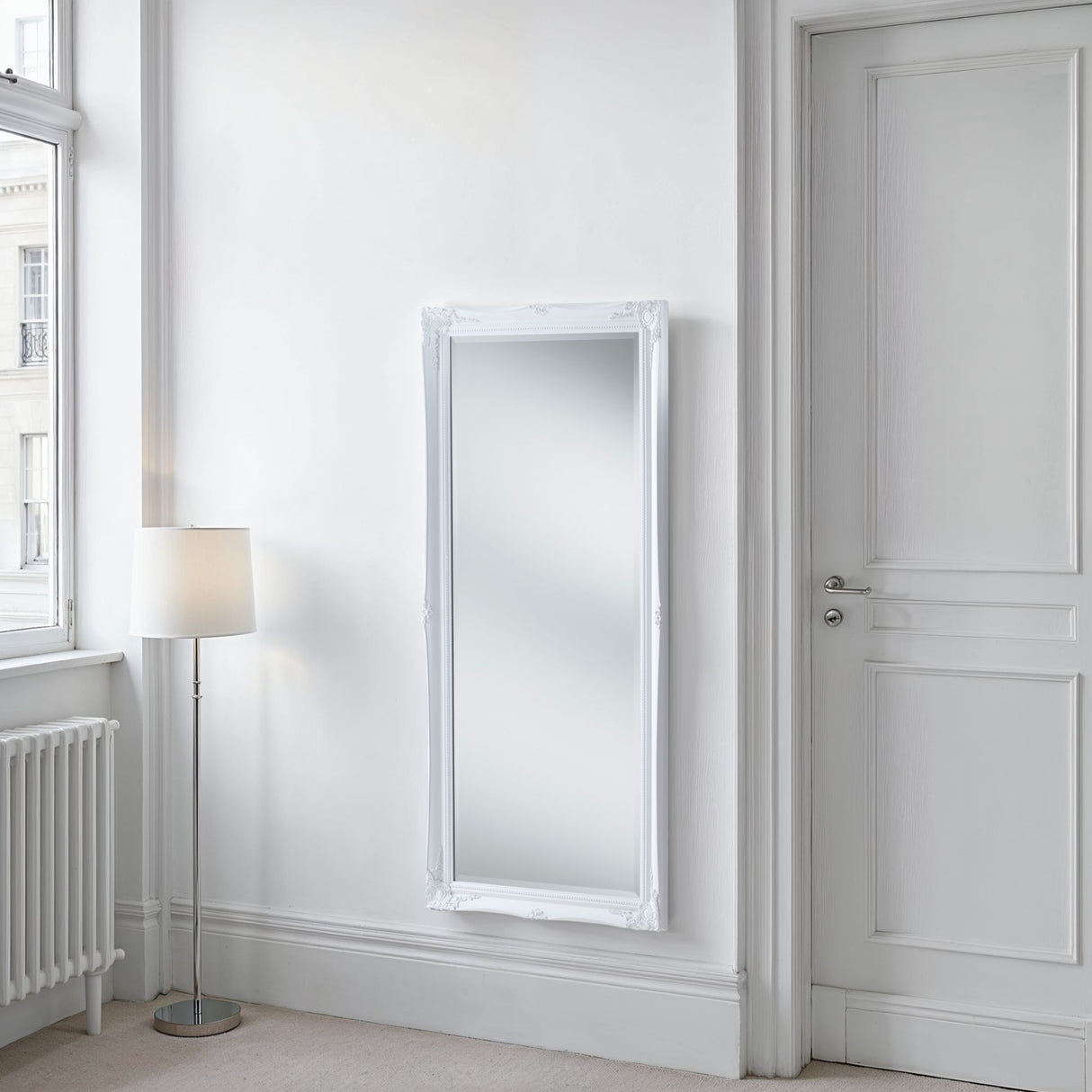 White wall mirror on a white wall next to a door and floor lamp.