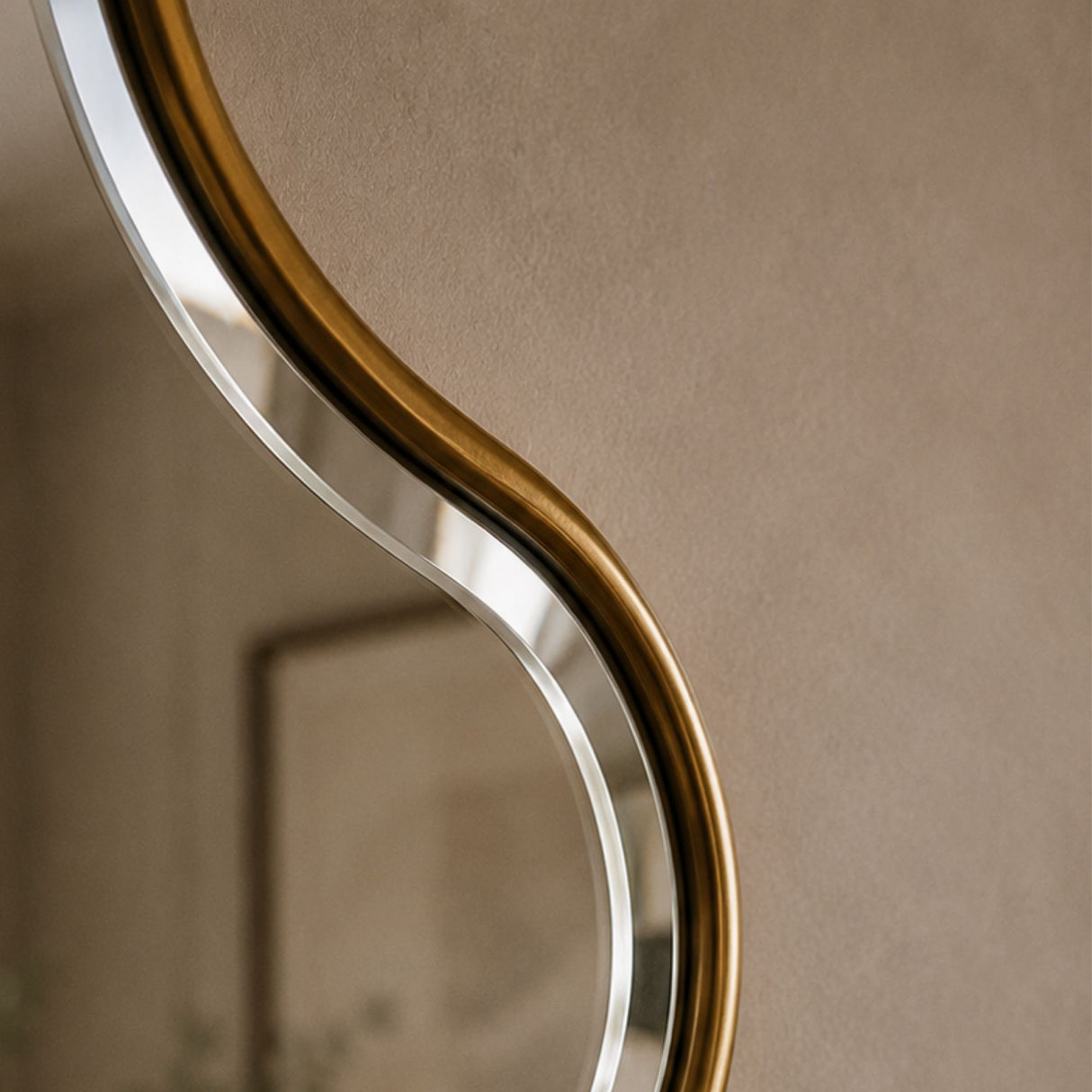 Close-up of a modern wall mirror with a flowing, wavy organic shape and a brushed gold metal frame against a warm beige wall.