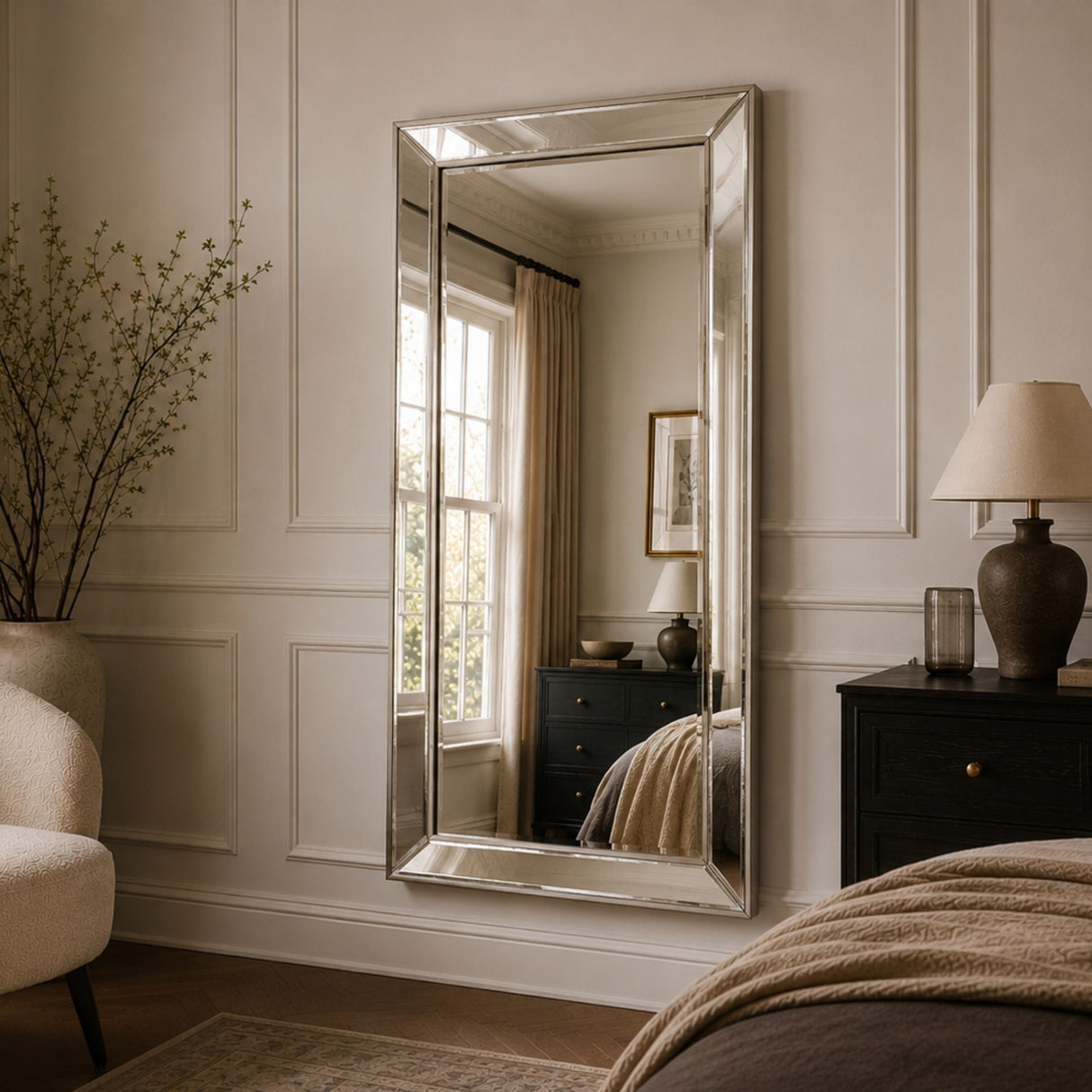 A tall, rectangular floor-to-ceiling mirror with a silver-toned frame hangs on a cream paneled wall in a styled bedroom. The mirror reflects a window and bed, while the room also features a dark wood nightstand, a table lamp, and a beige armchair.