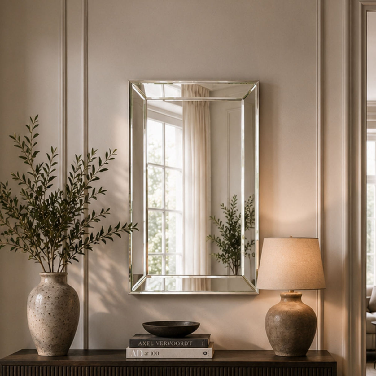 A rectangular wall mirror with a silver-toned frame hangs on a beige paneled wall above a console table. The scene is styled with a large textured vase of greenery on the left, a lit table lamp on the right, and books in the center.