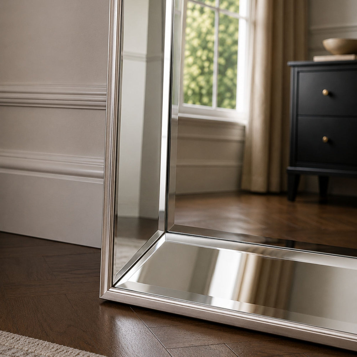A tall floor mirror with a slim silver metal frame standing on a dark wood floor in a styled room, reflecting a window and a black dresser.