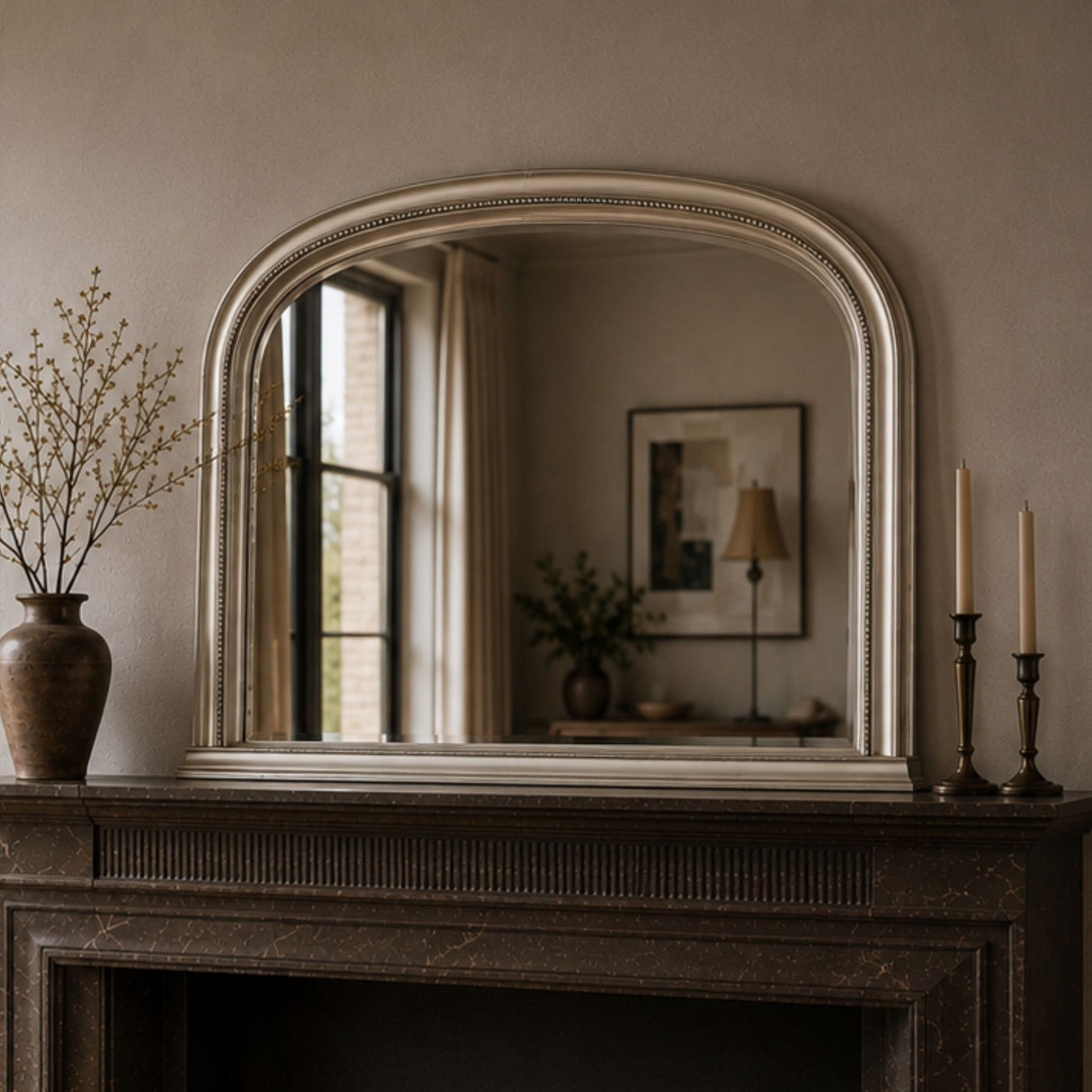 An arched wall mirror with a distressed silver frame resting on a dark fireplace mantel in a styled room. The mirror reflects a softly lit interior window and lamp, while the mantel is decorated with a brown ceramic vase containing dried branches on the left and two dark brass candlesticks with cream candles on the right.