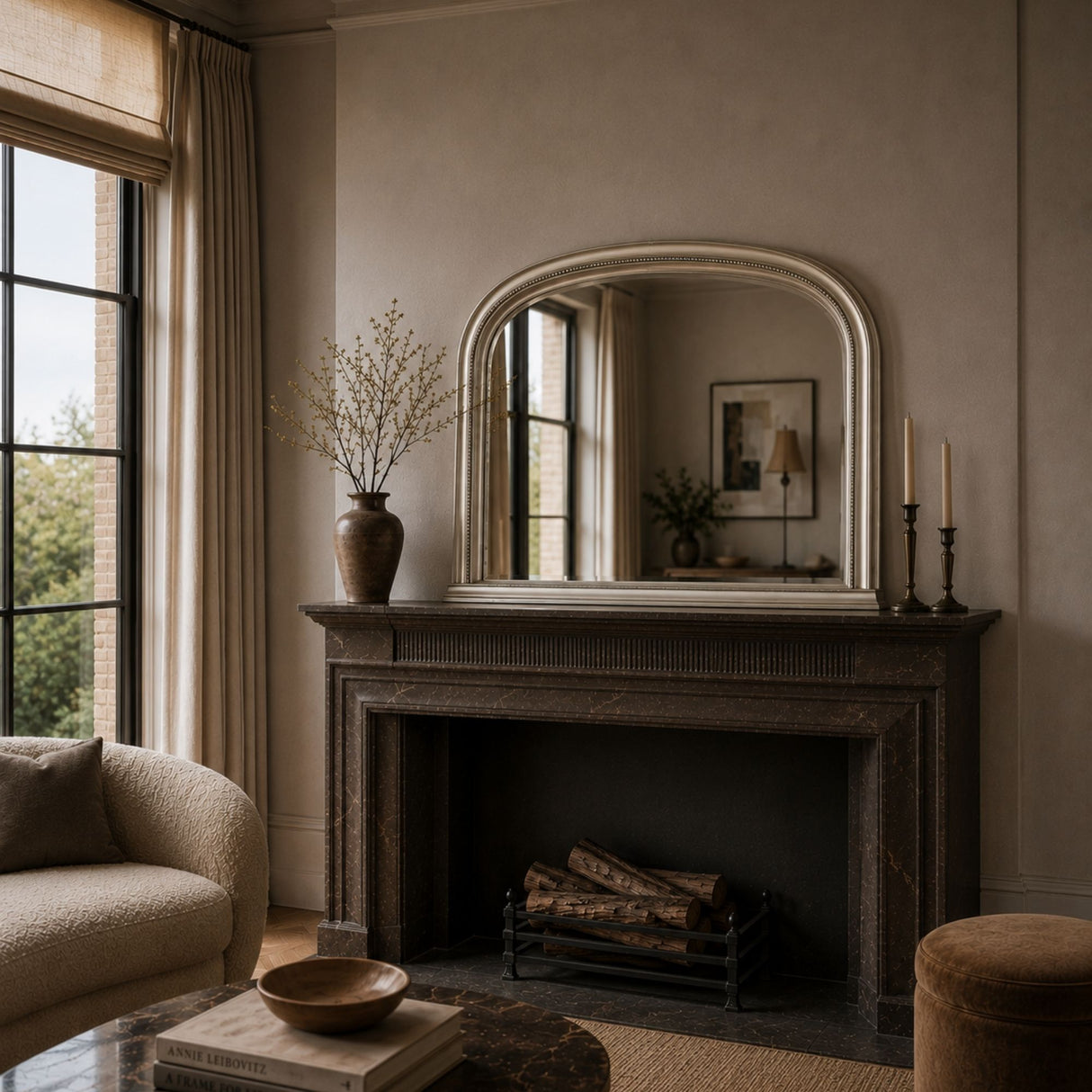 A large arched mirror with a cream frame hangs above a dark fireplace mantel in a styled living room. The mantel holds a tall vase with dried branches and candlesticks, and the room has warm neutral walls, curtains, and a sofa.