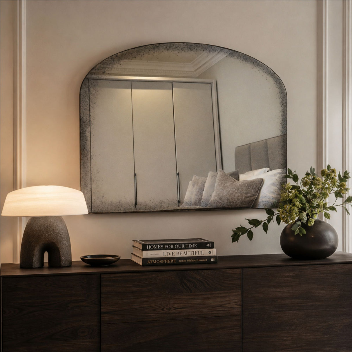 An arched rectangular wall mirror with a distressed, textured dark gray frame hangs above a dark wood console table in a styled bedroom setting. The mirror reflects a bed with gray pillows, and the room is illuminated by a lit table lamp on the left and natural light from the right.