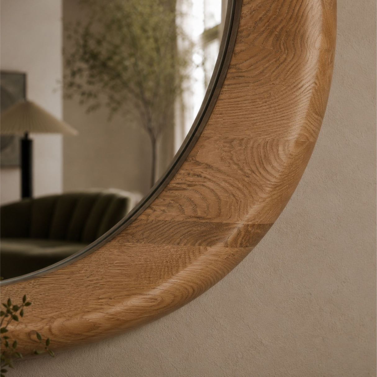 A close-up of a large oval wall mirror with a thick, curved natural oak wood frame, mounted on a textured beige wall. The reflection in the mirror shows a softly lit living room interior with a green sofa and a lamp.