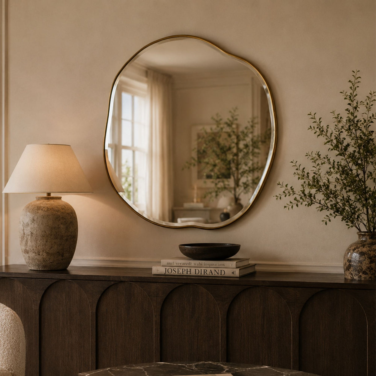 An organic-shaped round wall mirror with a gold-toned frame hangs above a dark wood console table in a warm, neutral-toned room. The mirror reflects a softly lit interior window, while the table is styled with a textured ceramic table lamp, a black bowl, a book, and a vase with dried branches.