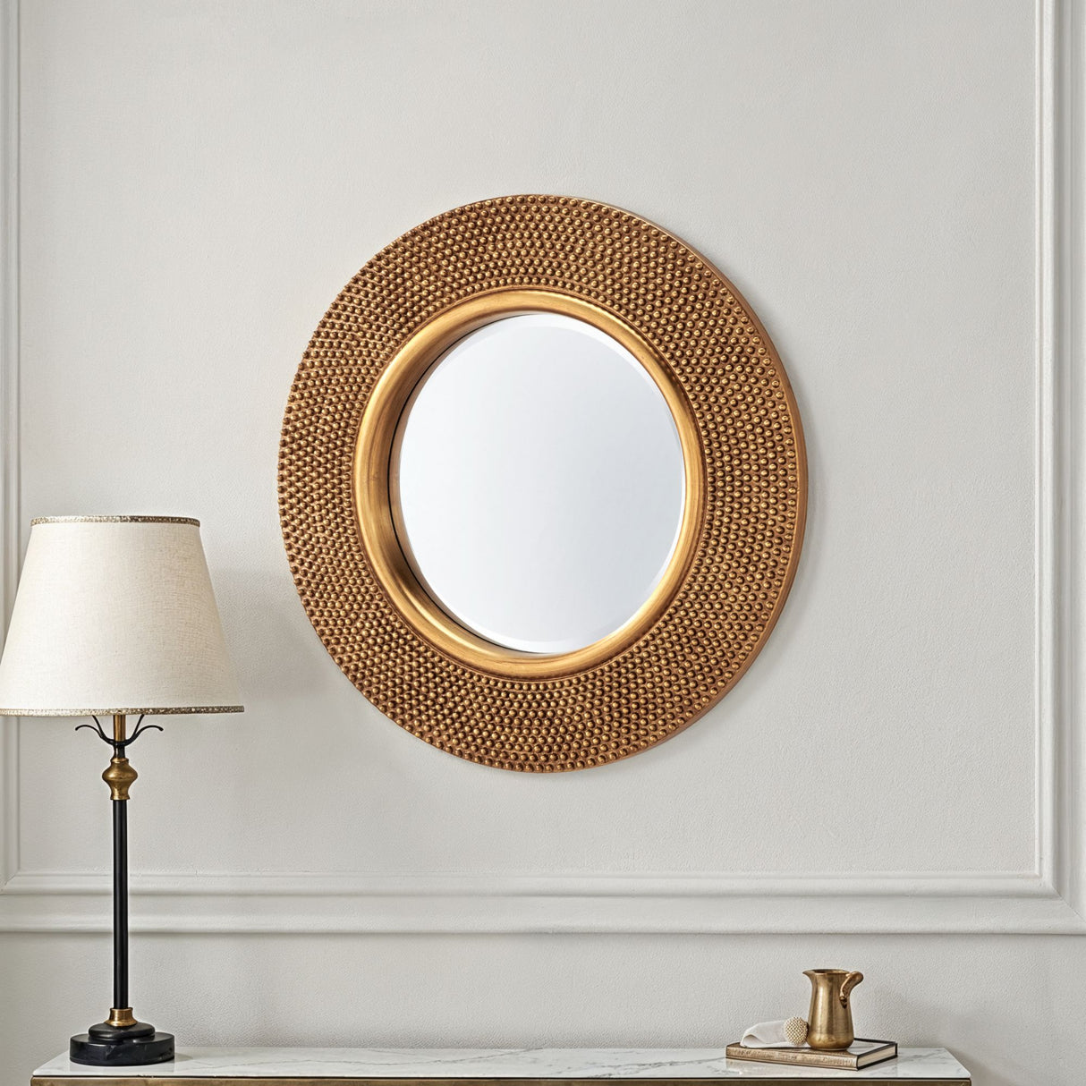 Round gold-framed mirror on a wall with a lamp and decorative items below.
