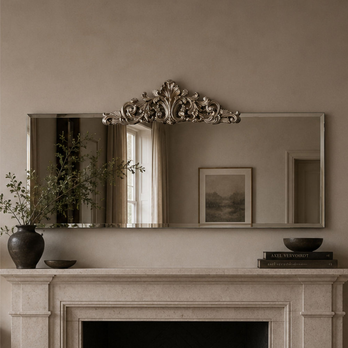 An ornate rectangular wall mirror with a decorative silver-toned crest at the top, mounted above a fireplace mantel in a warm neutral room. The mirror reflects a window and framed artwork, while the mantel holds a dark vase with branches, a small bowl, and books.