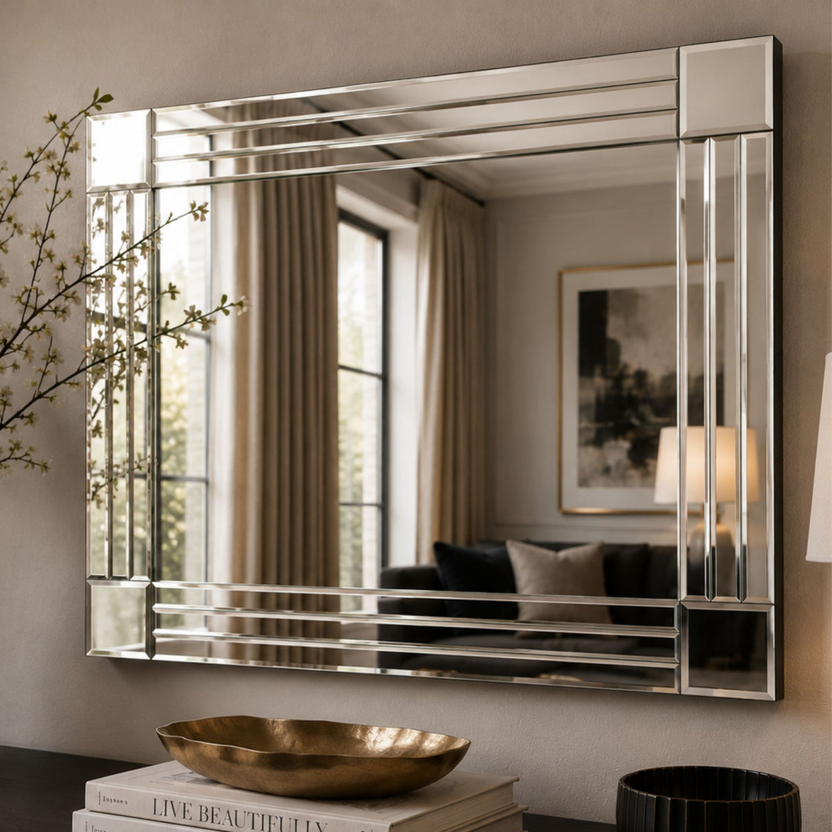 A large rectangular wall mirror with a polished silver frame and geometric segmented panel detailing, mounted on a beige wall. The mirror reflects a softly lit room with curtains, a sofa, and framed art, while books and a gold bowl sit below on a dark surface.