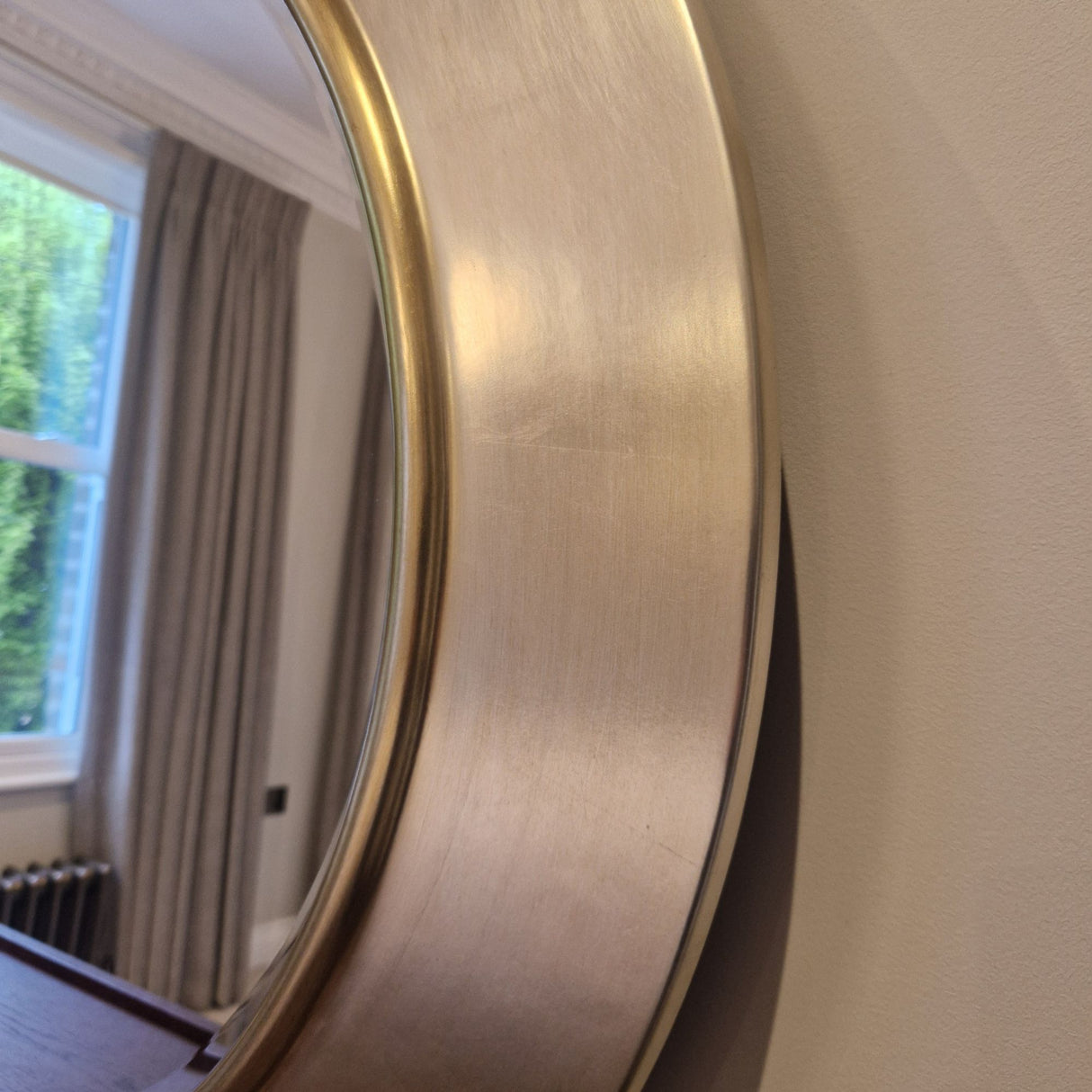Round gold-framed mirror on a beige wall with a window and curtains in the background.