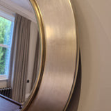 Round gold-framed mirror on a beige wall with a window and curtains in the background.