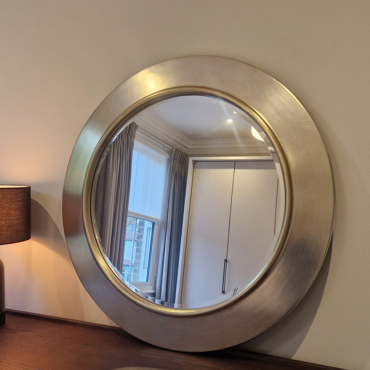 Round metallic wall mirror on a neutral wall with a lamp and window reflection.