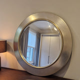 Round metallic wall mirror on a neutral wall with a lamp and window reflection.