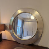 Round silver-framed mirror on a wall with a lamp and window reflection.