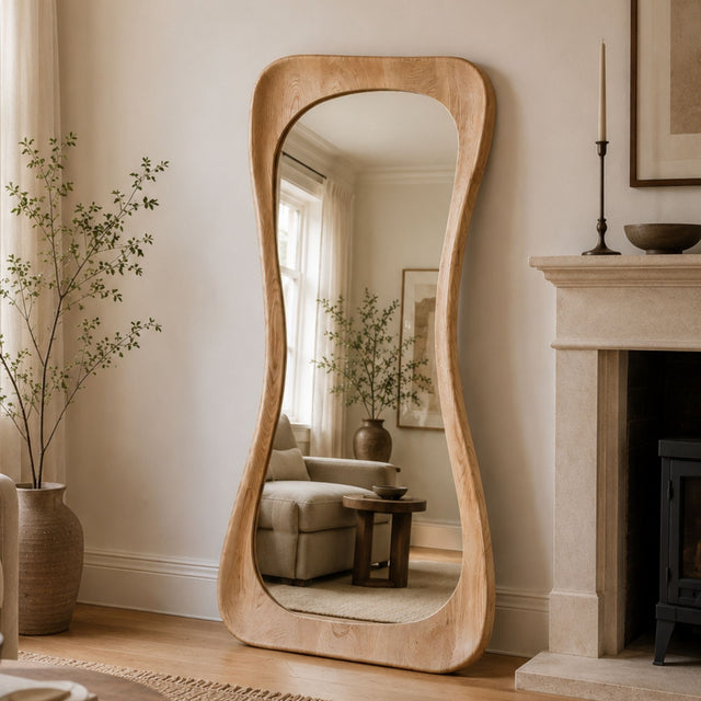 A tall, full-length floor mirror with a wavy, organic light oak wood frame stands in a cozy, neutral-toned living room next to a fireplace and a large vase with dried branches