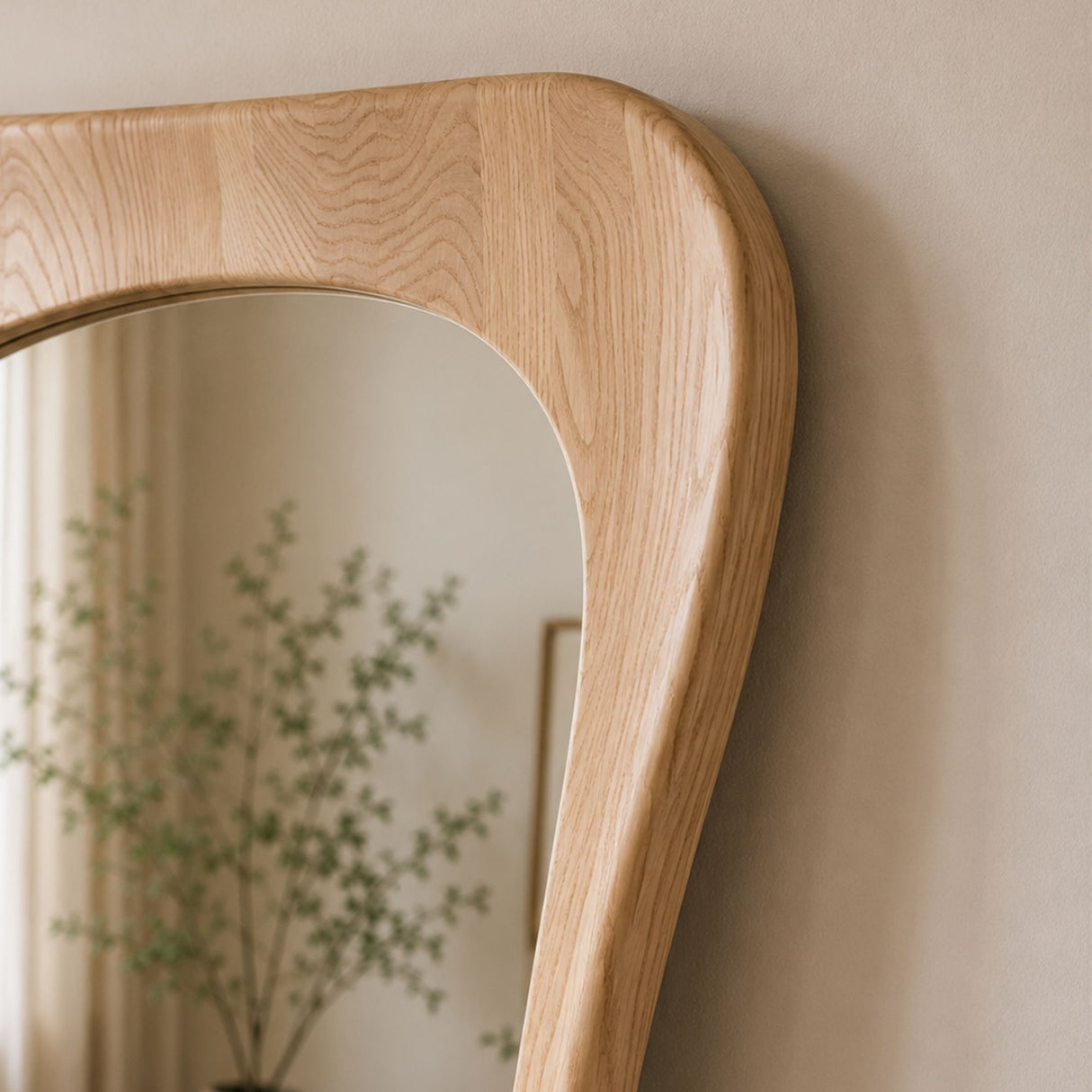 Close-up of a natural oak wood wall mirror with a curved, rounded top frame, leaning against a beige wall with its reflection showing a blurred plant.