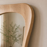 Close-up of a natural oak wood wall mirror with a curved, rounded top frame, leaning against a beige wall with its reflection showing a blurred plant.