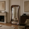 A tall floor mirror with a wavy, organic dark brown wooden frame stands in a styled living room next to a fireplace and upholstered armchair.
