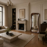 A styled living room interior with a cream fireplace, wood floors, a marble coffee table, upholstered seating, and a tall asymmetrical wooden floor mirror.