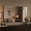 A large rectangular wall mirror with a slim silver frame hangs above a dark wood console table in a styled interior. The mirror reflects a softly lit room with curtains, a lamp, framed art, and cushions, while the room itself is lit in warm neutral tones.