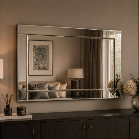A large rectangular wall mirror with a slim silver frame hangs above a dark wood console table in a styled interior. The mirror reflects a softly lit room with curtains, a lamp, framed art, and cushions, while the room itself is lit in warm neutral tones.