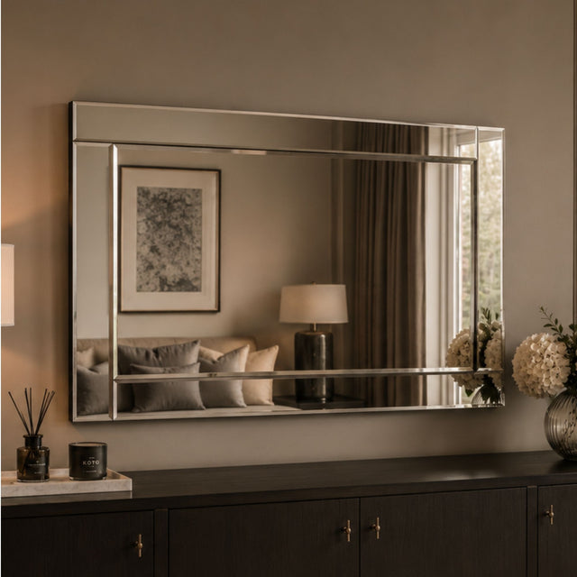 A large rectangular wall mirror with a slim silver frame hangs above a dark wood console table in a styled interior. The mirror reflects a softly lit room with curtains, a lamp, framed art, and cushions, while the room itself is lit in warm neutral tones.