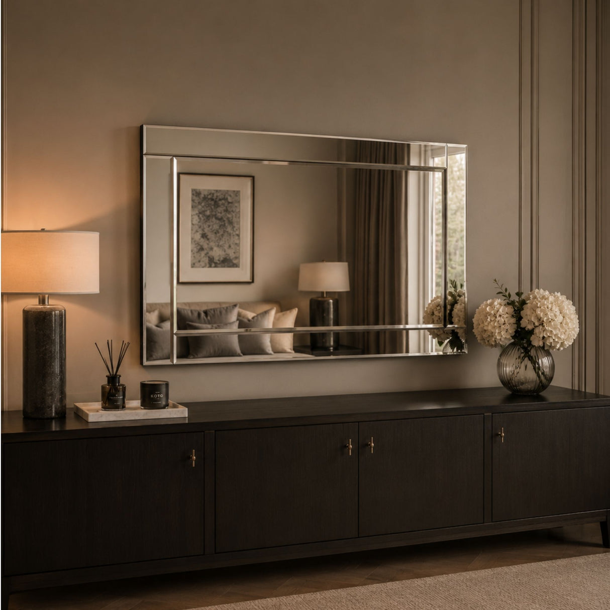 A large rectangular wall mirror with a thin metallic frame hangs above a dark wood console table in a styled interior. The room has warm neutral walls, a lit table lamp on the left, and a vase of white flowers on the right.