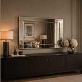 A large rectangular wall mirror with a thin metallic frame hangs above a dark wood console table in a styled interior. The room has warm neutral walls, a lit table lamp on the left, and a vase of white flowers on the right.