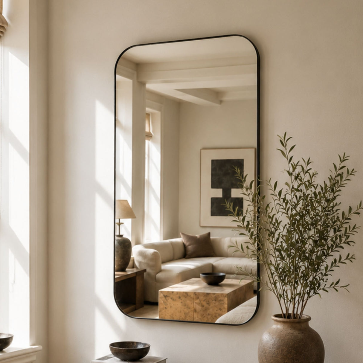 A tall, narrow rectangular wall mirror with a thin black frame hangs on a warm beige wall in a sunlit room. The mirror reflects a softly lit living room with a cream sofa, wood table, and framed art.
