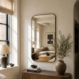 A tall, narrow wall mirror with a thin black frame hangs on a beige wall in a sunlit room. The mirror reflects a softly lit living room with a sofa and coffee table.