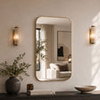 A large rounded rectangular wall mirror with a thin gold frame hangs on a beige wall, flanked by two matching gold wall sconces. The mirror reflects a softly lit living room interior, while the foreground shows a dark console table, a gray vase with greenery, a small amber candle, a book, and a ceramic bowl.