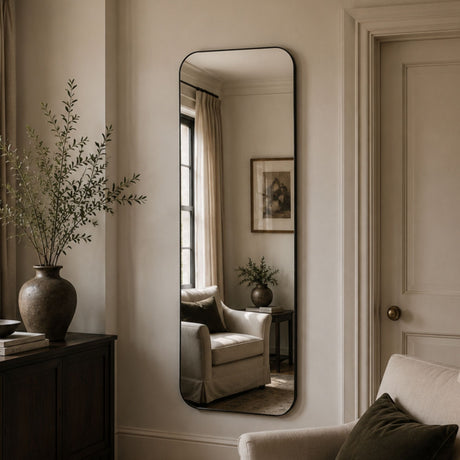 A tall, narrow floor mirror with a thin black frame hangs on a beige wall in a styled living room. The mirror reflects an armchair, side table, and framed artwork, while the room itself features a dark wood cabinet with a large vase of branches, a light armchair, and a paneled door.