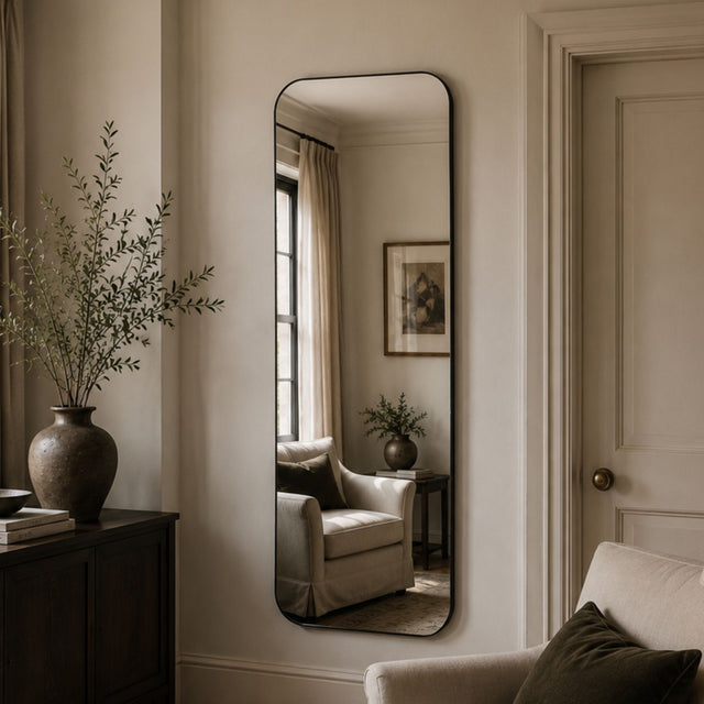 A tall, narrow floor mirror with a thin black frame hangs on a beige wall in a styled living room. The mirror reflects an armchair, side table, and framed artwork, while the room itself features a dark wood cabinet with a large vase of branches, a light armchair, and a paneled door.
