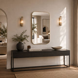 A rectangular wall mirror with a thin gold-toned frame hangs above a dark console table in a softly lit entryway. The mirror reflects part of a living room, while the room itself features warm beige walls, matching sconces, a vase with branches, books, and a bowl on the table.