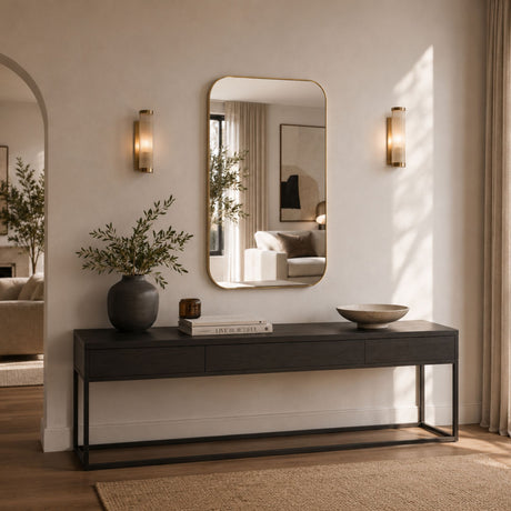 A rectangular wall mirror with a thin gold-toned frame hangs above a dark console table in a softly lit entryway. The mirror reflects part of a living room, while the room itself features warm beige walls, matching sconces, a vase with branches, books, and a bowl on the table.