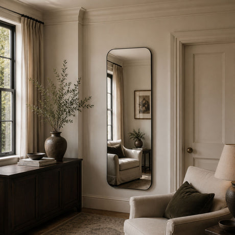 A tall, narrow floor mirror with a black frame hangs on a cream-colored wall in a styled living room. The mirror reflects a matching armchair and side table, while the room features a dark wood credenza, a large vase with dried branches, and a light beige armchair in the foreground.