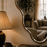 A wavy, organic-shaped wall mirror with a brushed silver metal frame and a gold-toned inner border, styled in a warm, cozy living room next to a lit table lamp.