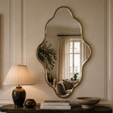 An ornate scalloped wall mirror with a gold-toned frame hangs on a cream paneled wall above a dark console table. A lit table lamp, books, and decorative bowls are visible in the room reflected in the mirror.