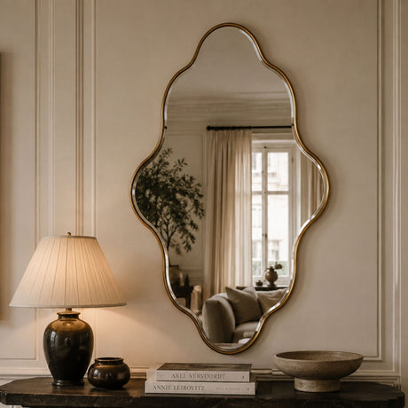 An ornate scalloped wall mirror with a gold-toned frame hangs on a cream paneled wall above a dark console table. A lit table lamp, books, and decorative bowls are visible in the room reflected in the mirror.