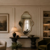 An ornate scalloped wall mirror with a gold-toned frame hangs on a cream paneled wall above a dark console table in a styled living room setting.