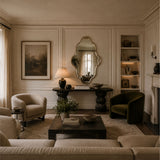 A styled living room interior with cream upholstered seating, a dark wood coffee table, a green velvet armchair, a console table with an ornate mirror, built-in shelving, and warm ambient lighting.