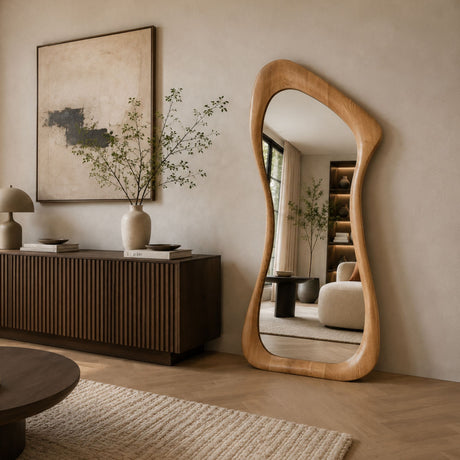 An organic-shaped floor mirror with a natural light wood frame stands in a modern, neutral-toned living room next to a dark wood credenza and a large abstract painting.
