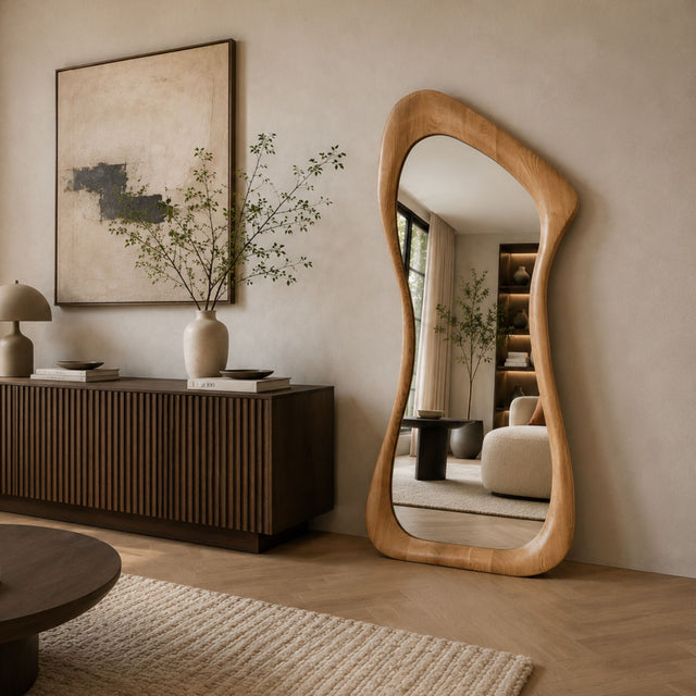 An organic-shaped floor mirror with a natural light wood frame stands in a modern, neutral-toned living room next to a dark wood credenza and a large abstract painting.