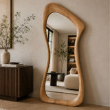 An organic-shaped wooden floor mirror with a natural light oak frame, leaning against a beige wall in a modern, minimalist living room setting.