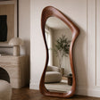 An organic-shaped full-length floor mirror with a rich, wavy brown wooden frame, styled in a warm, modern living room setting.